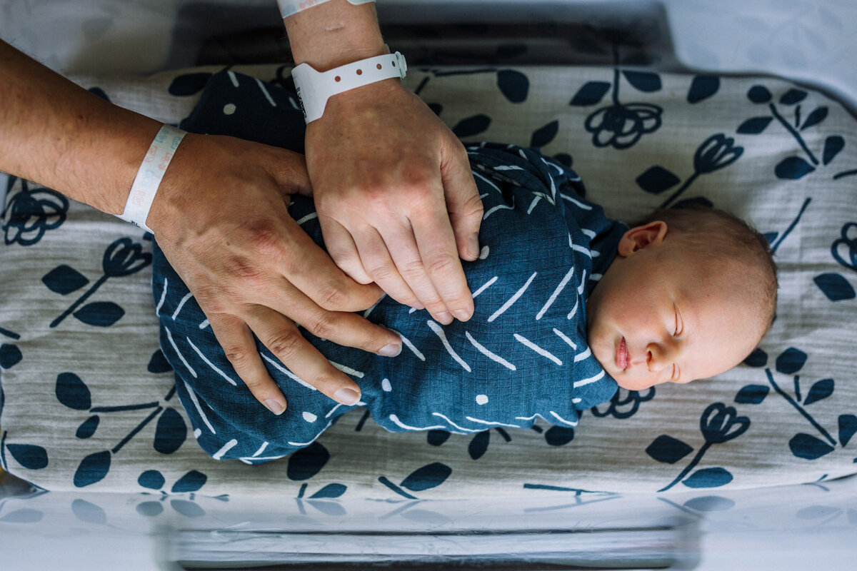 newborn sleeping in hospital bassinet with complimentary blue patterned blankets with mom and dad's hands with hospital bracelets on resting hands on baby