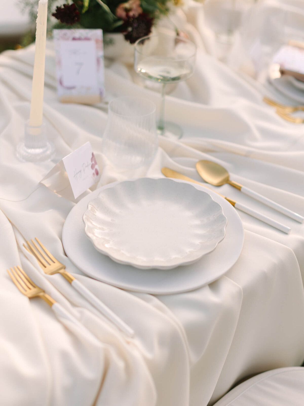 Elegant table setting with a white scalloped plate on a satin cloth, gold cutlery, a white candle, and floral centerpiece.