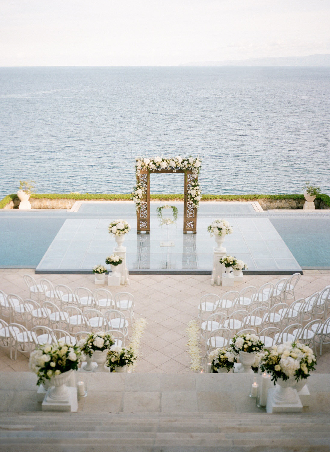 destination-wedding-bali-amankila-ceremony-over-pool-platform--altar-white-chairs-floral-strucutre