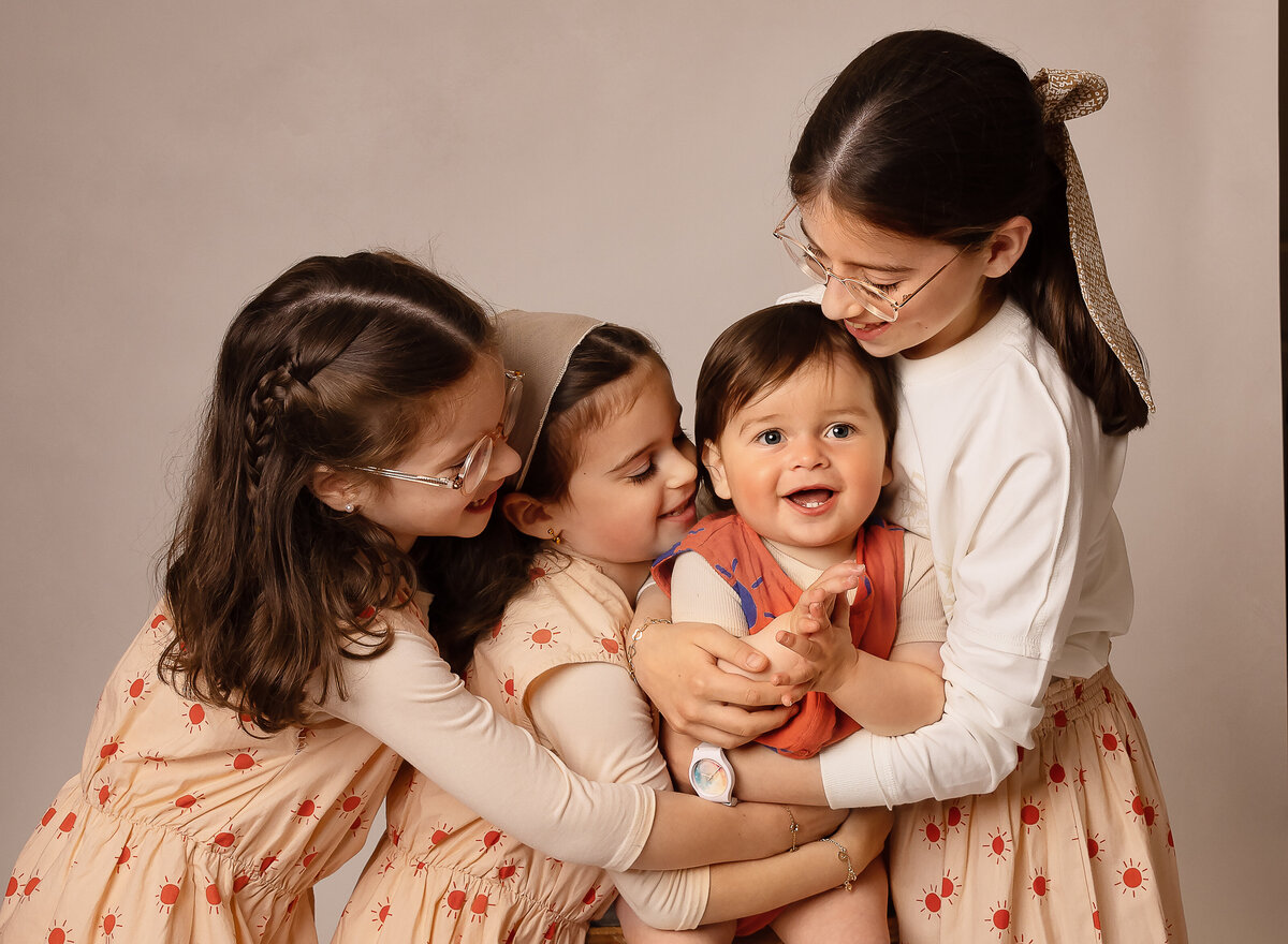 Studio portrait. Baby brother is being cuddled by his three big sisters. Baby boy is in the middle and the sisters are all looking at their little brother. Captured by best Brooklyn, NY family photographer Chaya Bornstein.