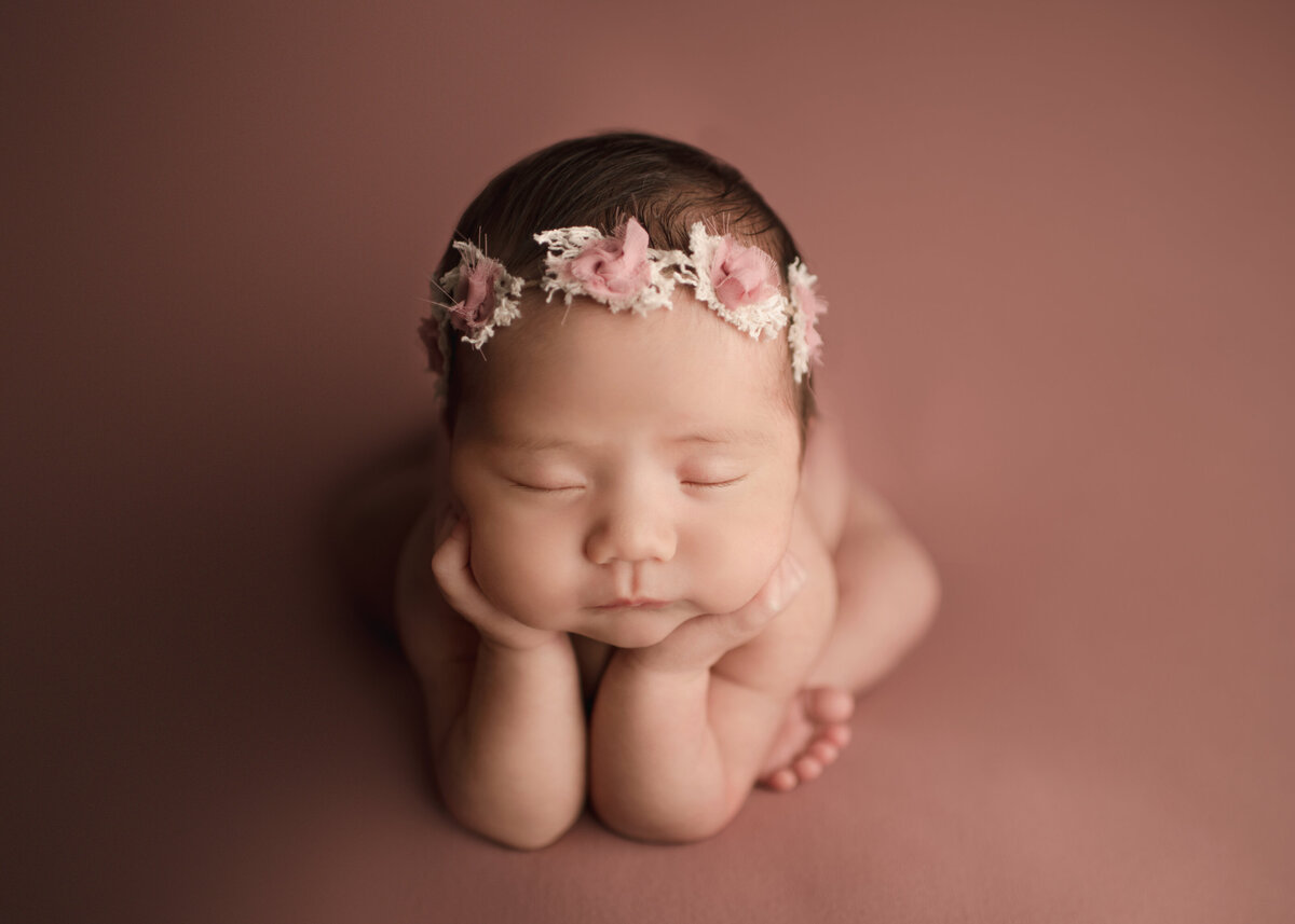 Newborn baby girl in froggy pose has her hands propped underneath her cheeks at her Lake Elsinore newborn photoshoot.
