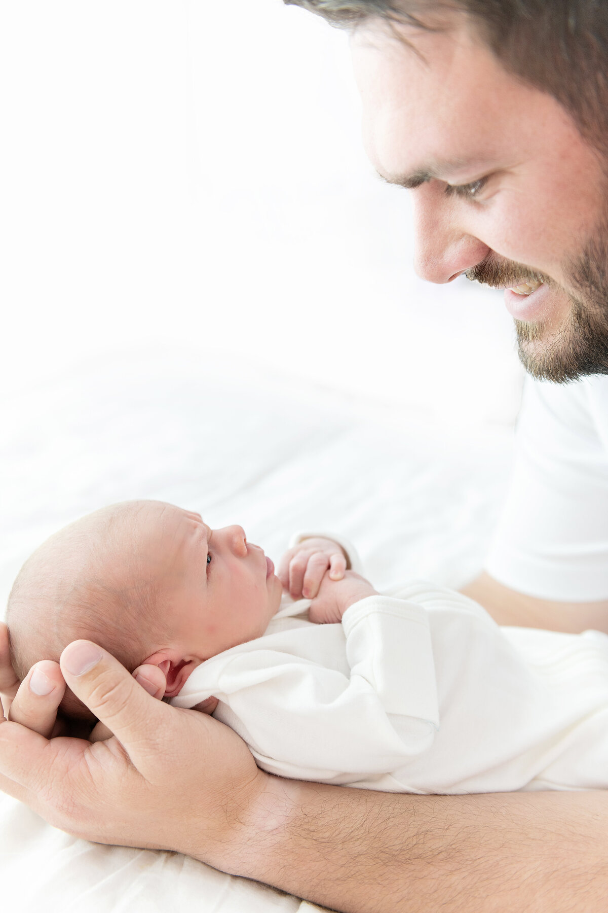 A happy new dad smiles down at his newborn baby in a white onesie in his hands