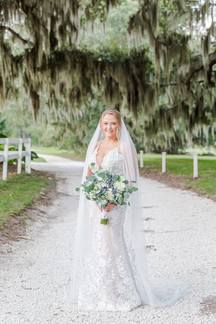 Beautiful bride stands alone in her dress and veil on a dirt path with her bouquet in front of large Oak trees and hanging moss.