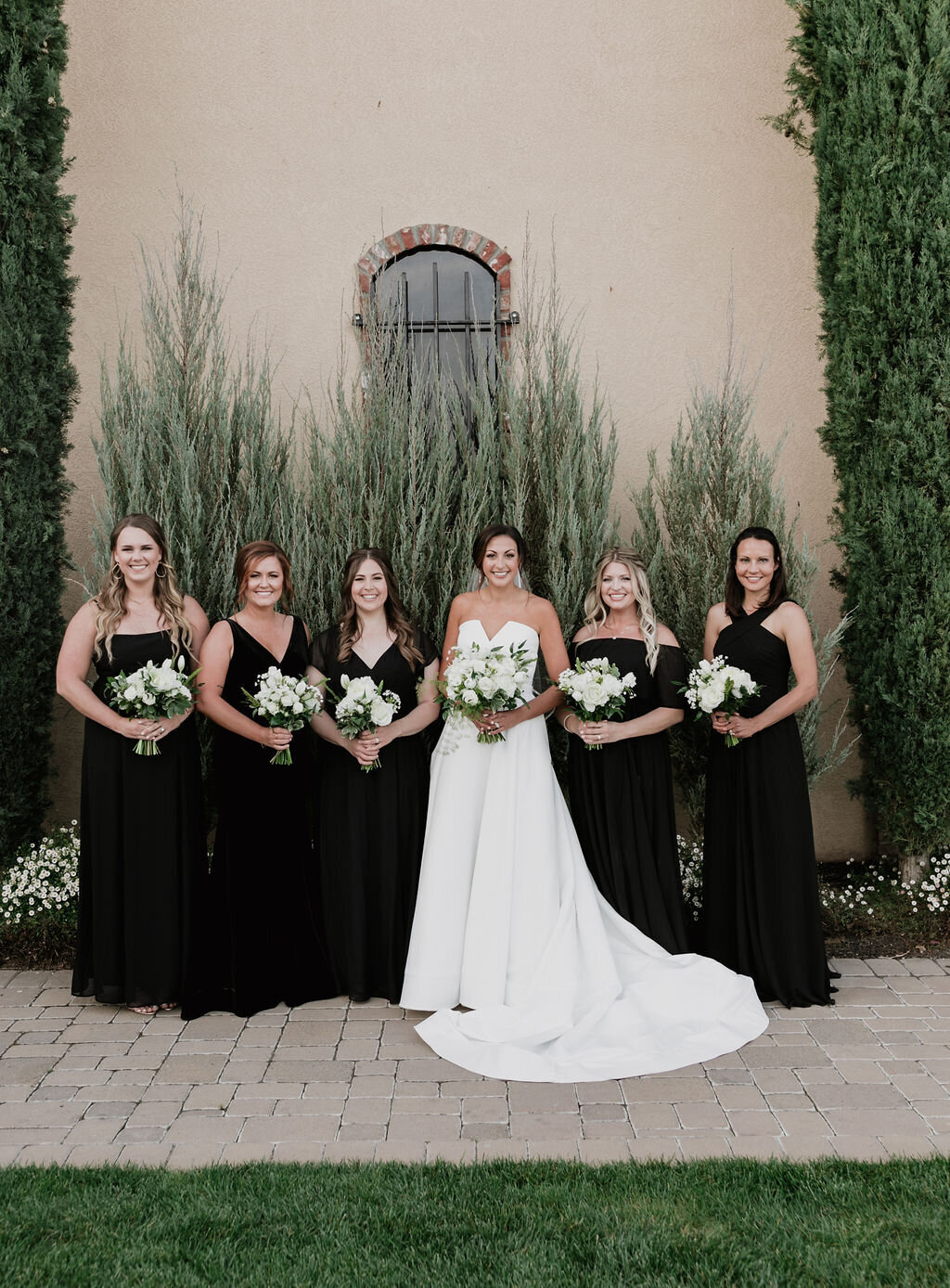 Bridal party at the Palm Event Center in Pleasanton, CA, during a spring wedding. The bride and her bridesmaids, dressed in elegant black satin gowns, hold white flower bouquets