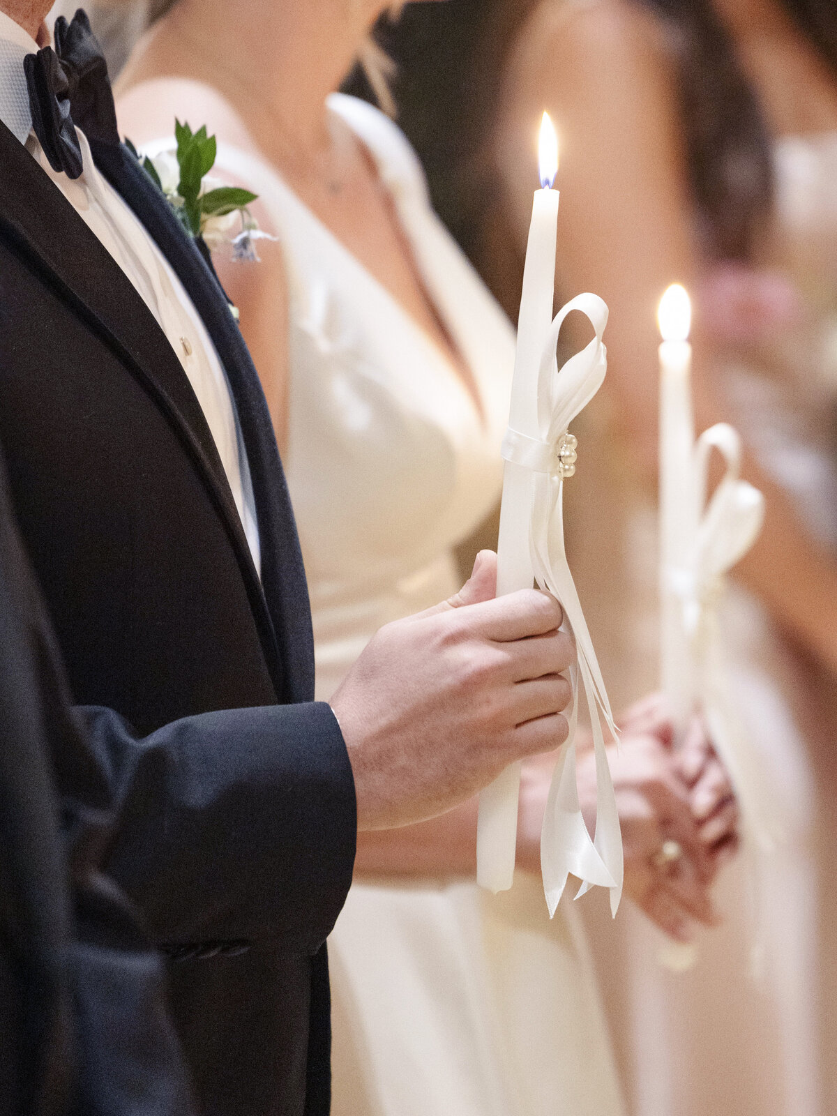 People dressed formally hold lit white candles with ribbons during a ceremony. The focus is on the hands holding the candles and the soft glow of the flames.