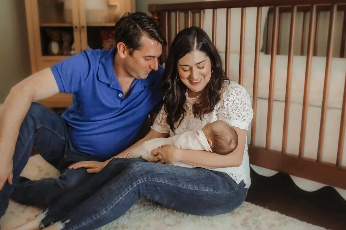 A man and woman sit on the floor by a crib, smiling down at a swaddled baby in the woman's arms during a Macon in home newborn photography session by Fire Family Photography.