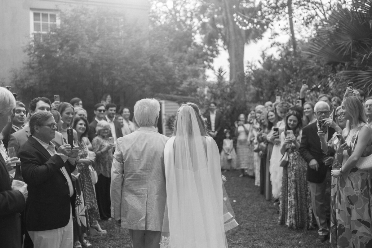 Black and white photo of bride walking down the aisle with her father during a backyard wedding ceremony in Charleston, South Carolina.