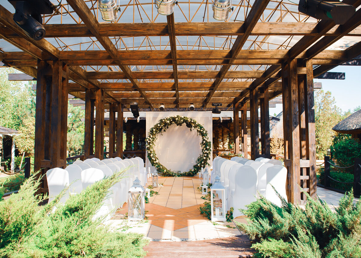 A wedding ceremony is set in a garden, under a pergola with a circular ceremony arch of greenery.
