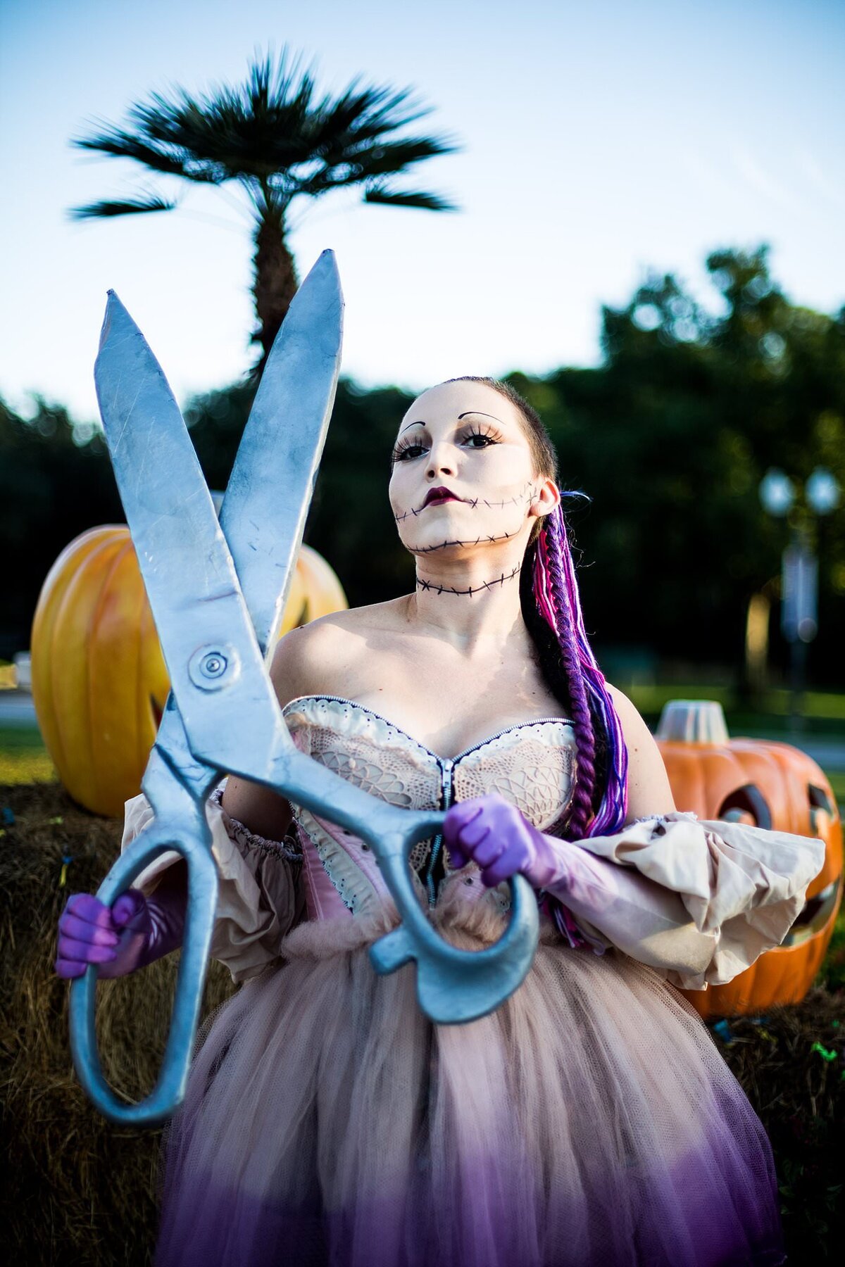 Halloween stitched doll costume and make up photo, the woman is holding a huge pair of scissors and she's among pumpkins looking creepy