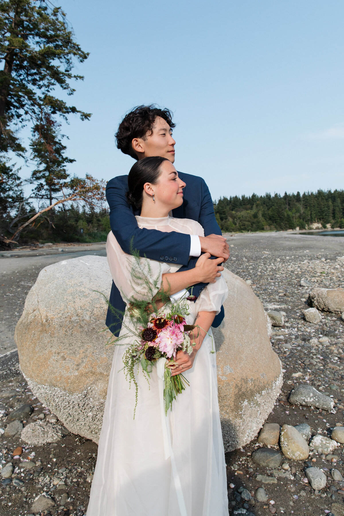 Groom wraps his arms around the bride in front of a large rock on a Puget Sound Beach on their wedding day.