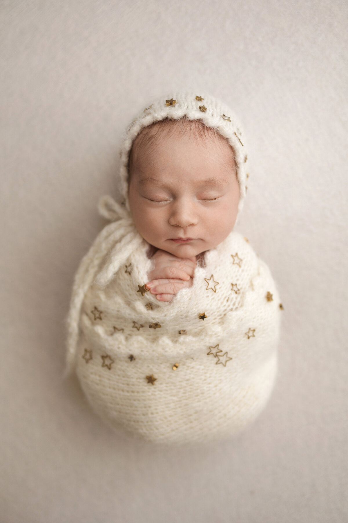 Newborn baby wrapped in a white knit blanket adorned with golden stars, sleeping peacefully with a matching white knit bonnet.