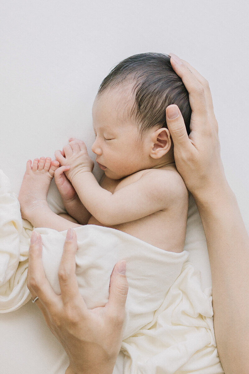 Baby sleeping on his side with his legs curled up. The mother is holding baby's head and bottom. Shot in Yokohama by newborn photographer Yukika Ishikawa Photography
