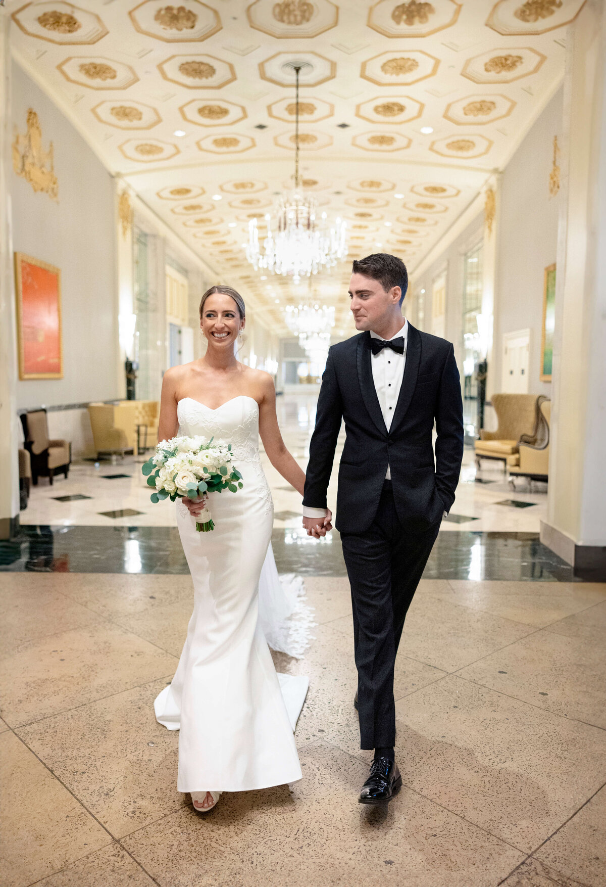 A bride and groom walk hand in hand down an elegant hallway with chandeliers. The bride wears a strapless white gown and carries a bouquet of white flowers, while the groom is in a black tuxedo. Both are smiling.
