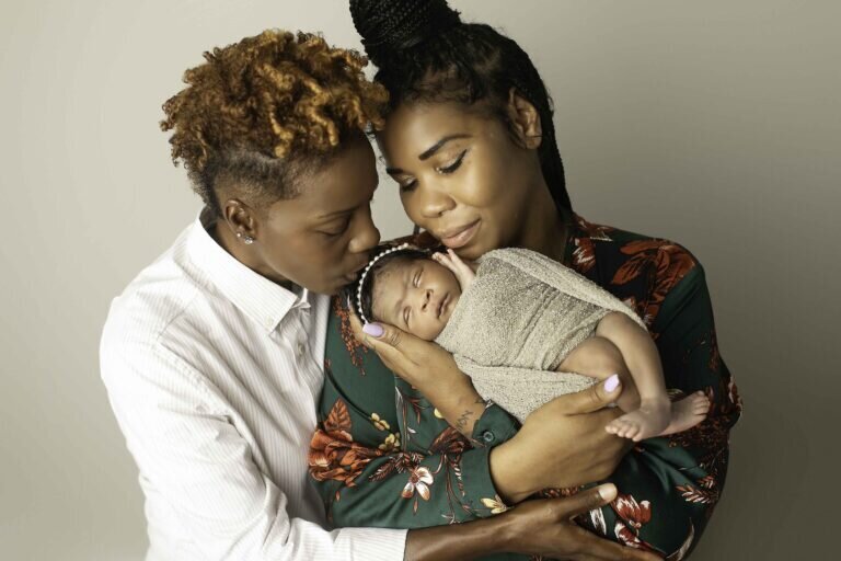 Two loving moms snuggling close and sharing a sweet kiss while holding their wrapped newborn baby, capturing a heartwarming and intimate family moment
