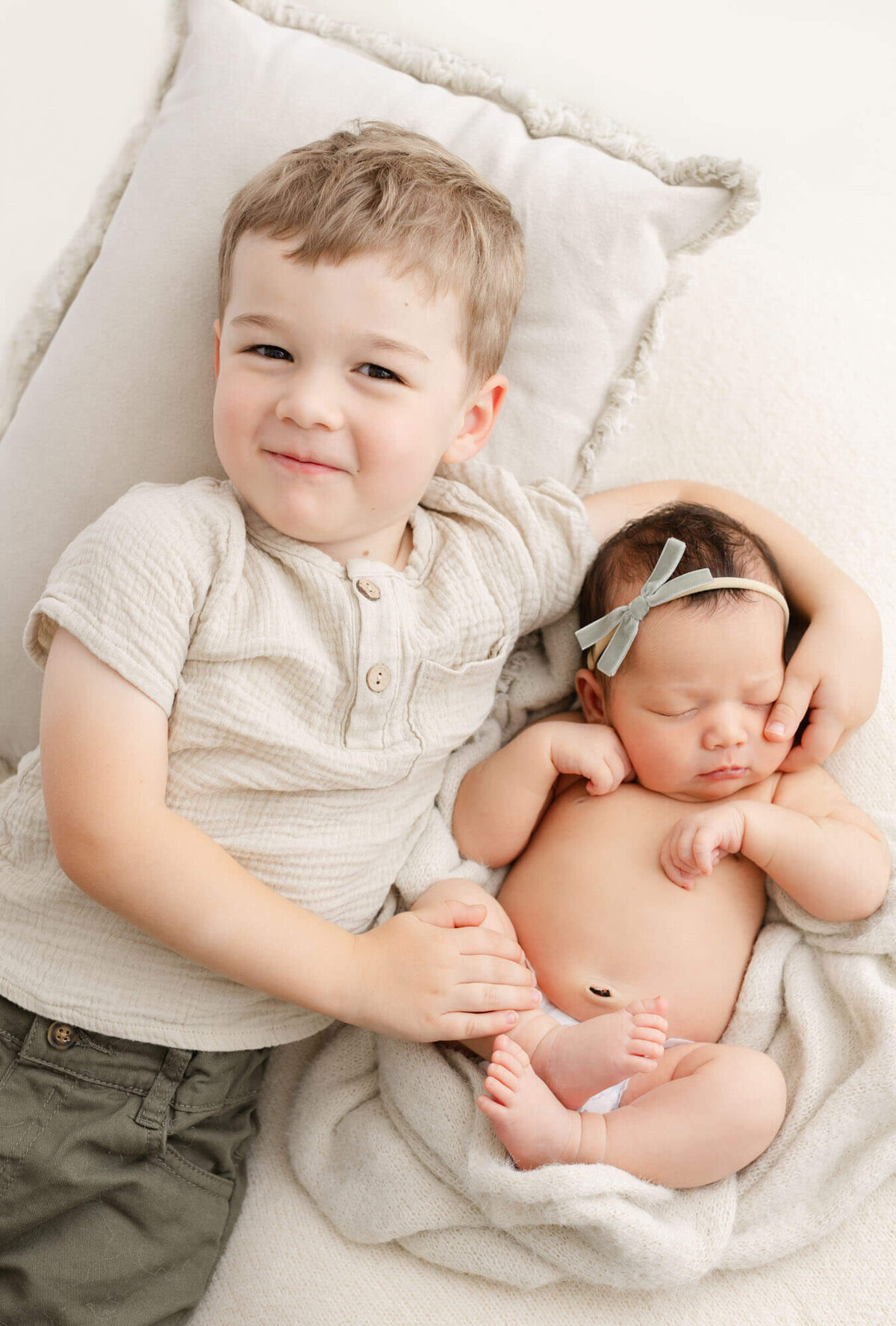 Proud big brother holding his new baby sister while laying down next to her and brother is smiling up at camera. Baby is sleeping.