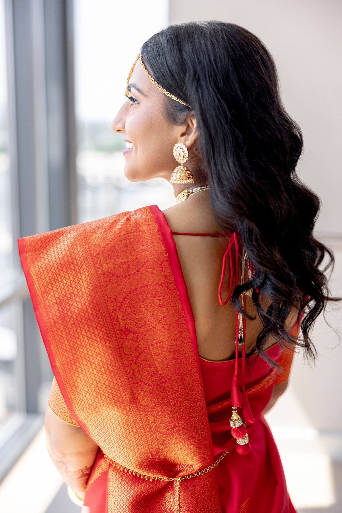 A woman in a red saree with gold patterns stands by a window, looking outside. She has long, dark hair and is wearing gold jewelry, including earrings and a headpiece. The scene is bright and serene.