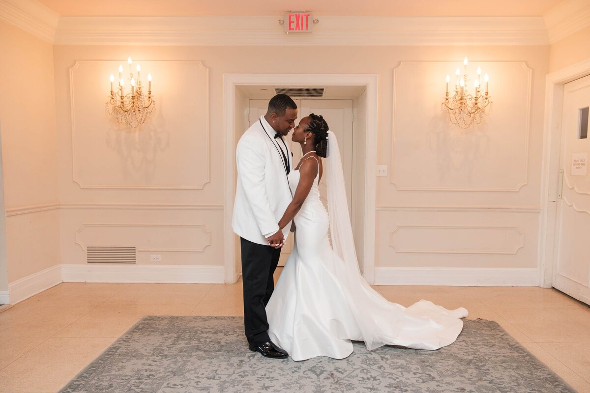 A bride and groom share a romantic moment, holding hands and smiling at each other inside the elegant interior of Azaleana Manor in Orange Park, Florida. The groom wears a classic white tuxedo jacket while the bride stuns in a sleek, fitted gown with a dramatic train and long veil. Set against a backdrop of ivory walls, chandeliers, and timeless details, this portrait captures the love and grace of their day. Photographed by Heartful Films, a Jacksonville wedding photographer and videographer known for timeless, elegant wedding imagery.