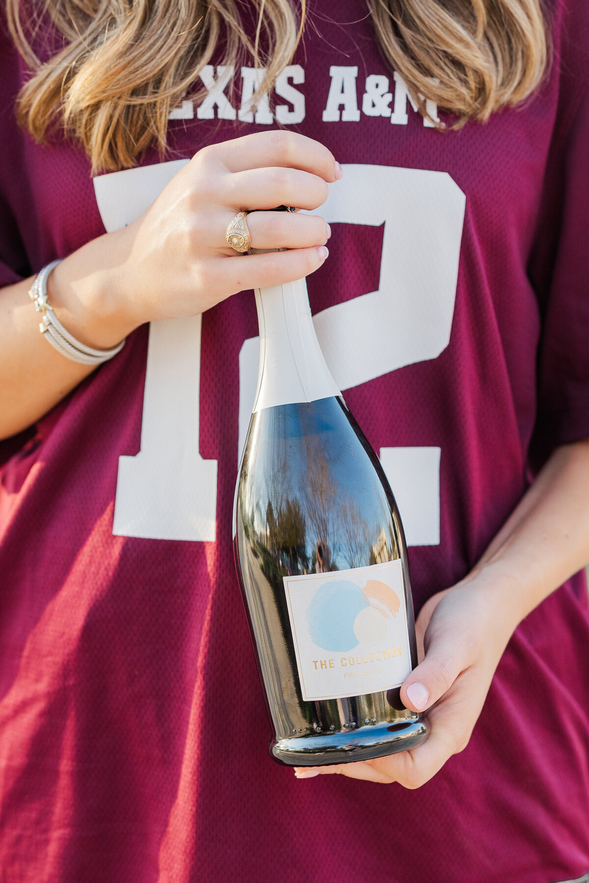 Close-up image of a Texas A&M senior holding a champagne bottle, featuring her Aggie ring and maroon jersey, captured in College Station, Texas.