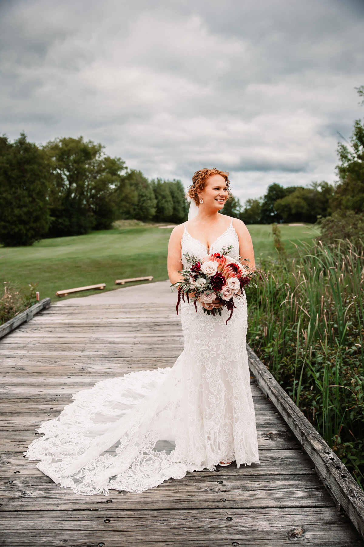 A bride in a long lace dress smiles while gazing out over a pond from a wooden bridge