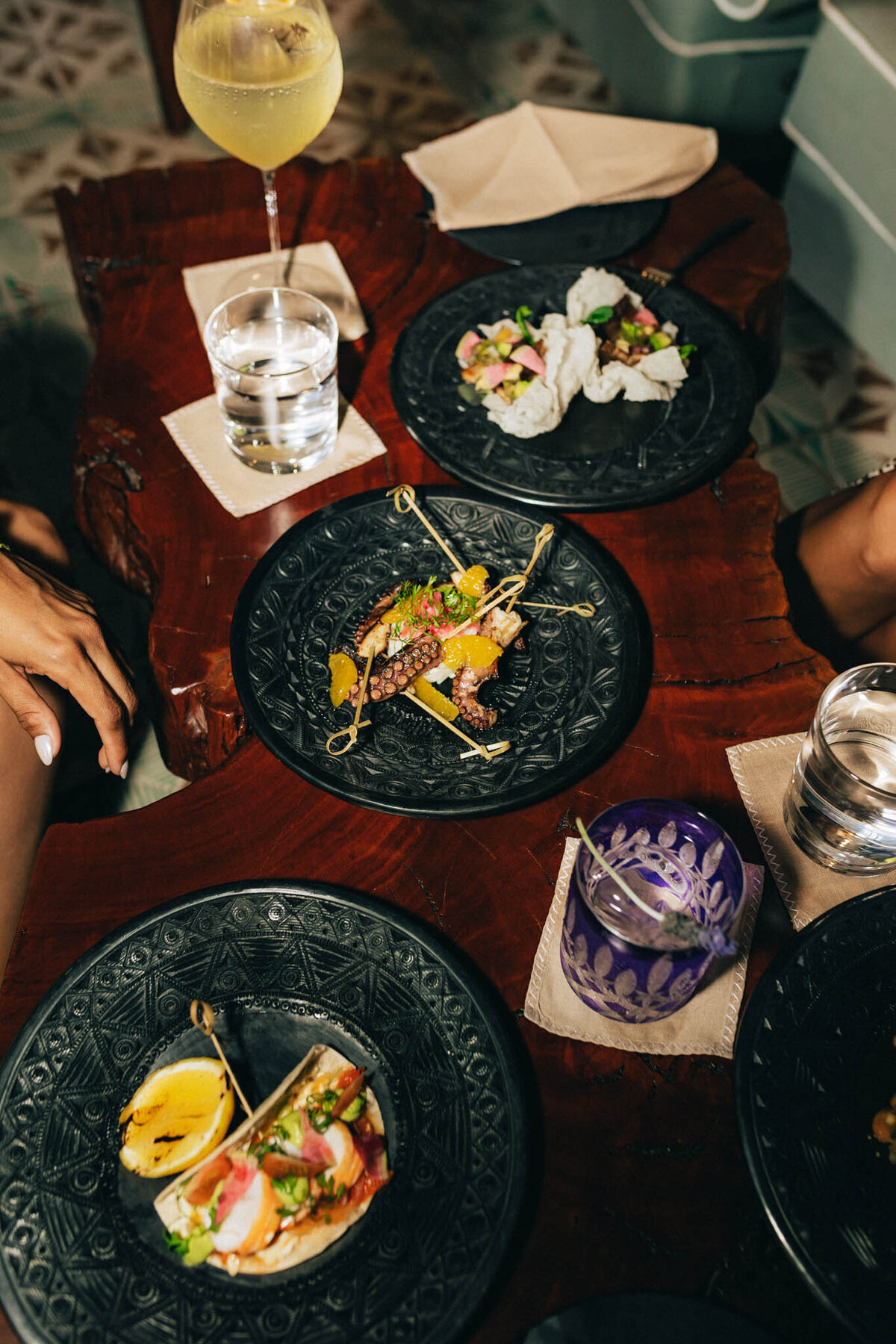 A wooden table with three black plates of gourmet food. One plate has tacos, another has a dish with skewered meat and vegetables, and the third has a seafood salad. Drinks include a yellow cocktail and glasses of water.