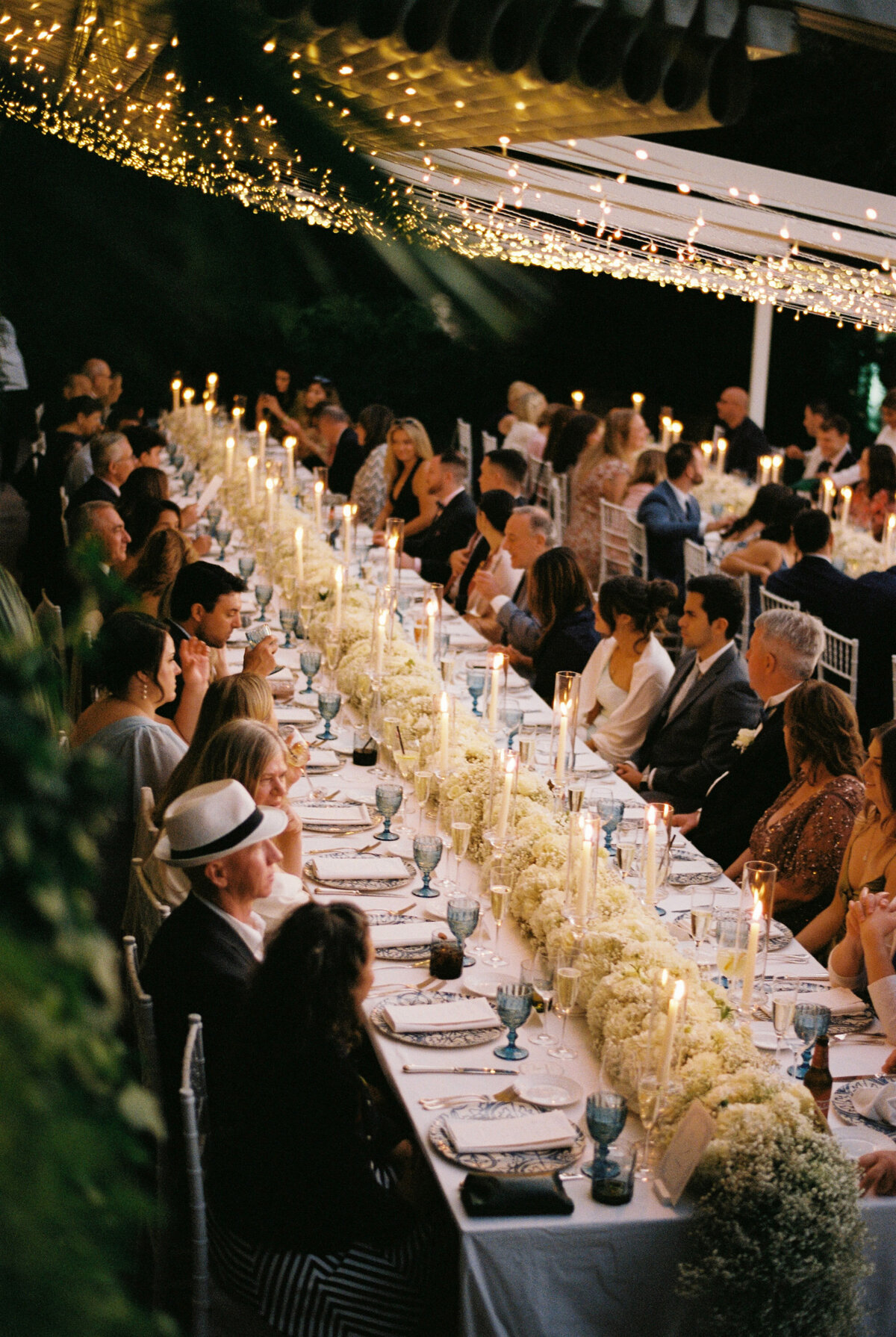 Elegant outdoor reception at Villa Silvana in Sorrento, with guests seated at a long, candlelit table adorned with white floral arrangements under a canopy of twinkling string lights, creating a romantic ambiance for a destination wedding.