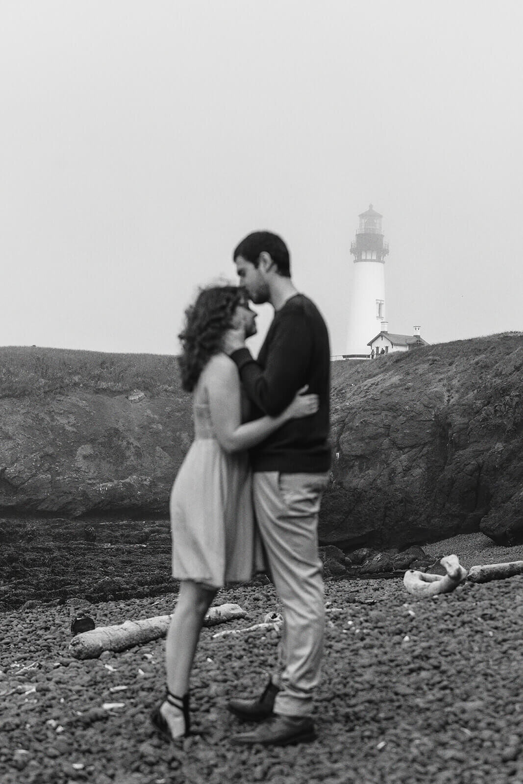 A couple embracing in black and white with a lighthouse in the distance behind them. 