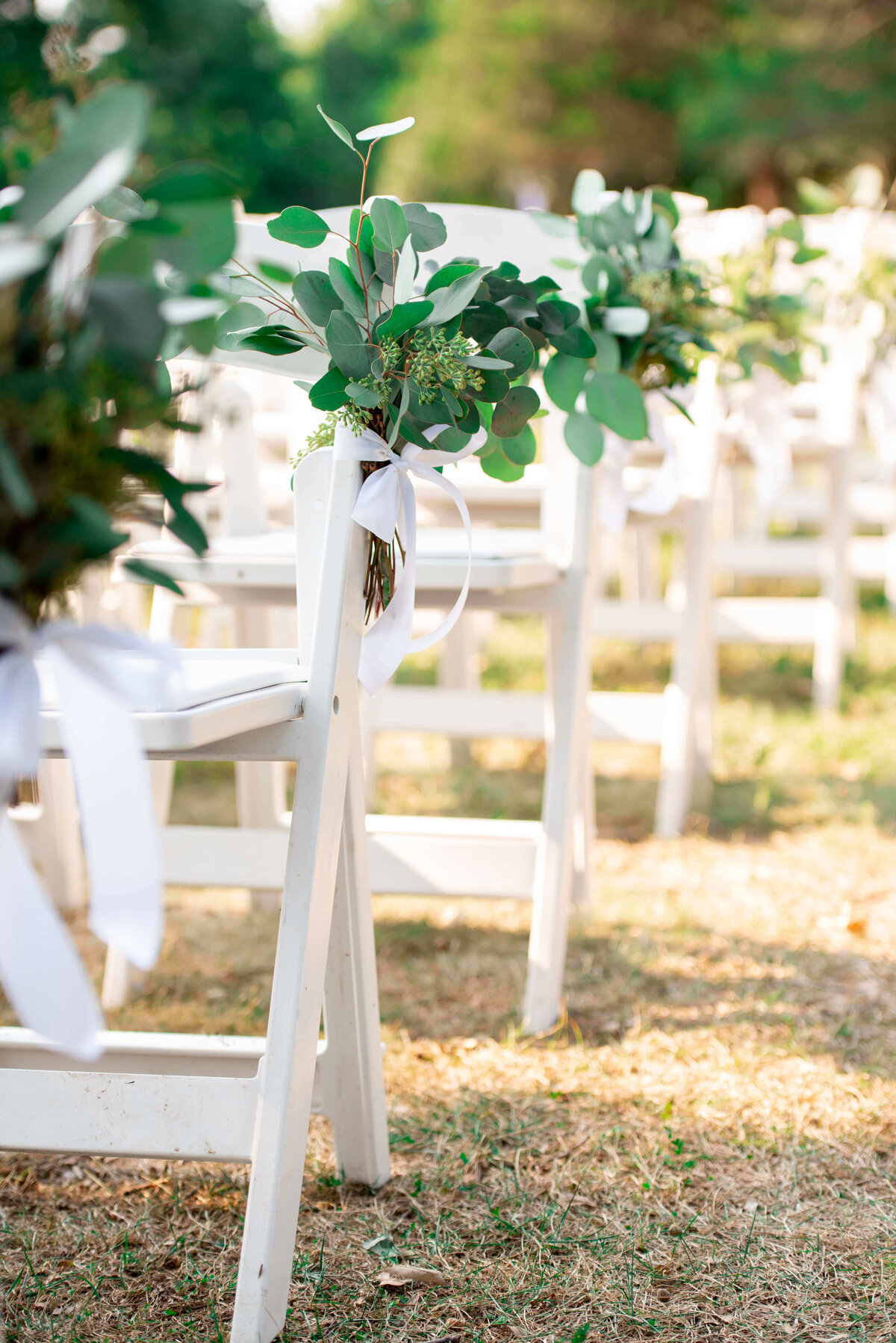 Rows of white chairs lined up outdoors, each decorated with green eucalyptus leaves and white ribbons. The chairs are set on a grassy area with blurred greenery in the background, suggesting a wedding or event setting.