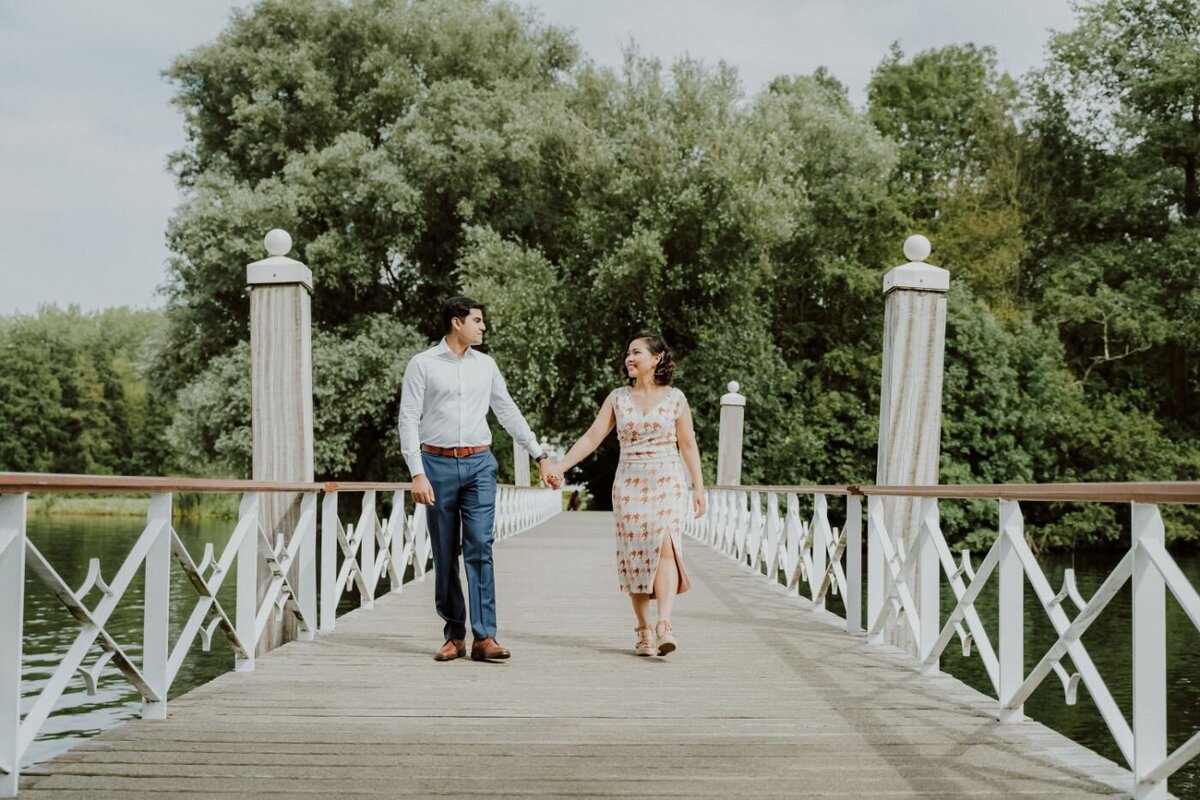 A vertical shot of a couple from behind, holding hands and standing on a path surrounded by trees. They are looking out over a body of water. The man is wearing a dark blue suit and the woman is wearing a white tiered dress. The sunlight filters through the dense foliage, creating a serene and intimate atmosphere.