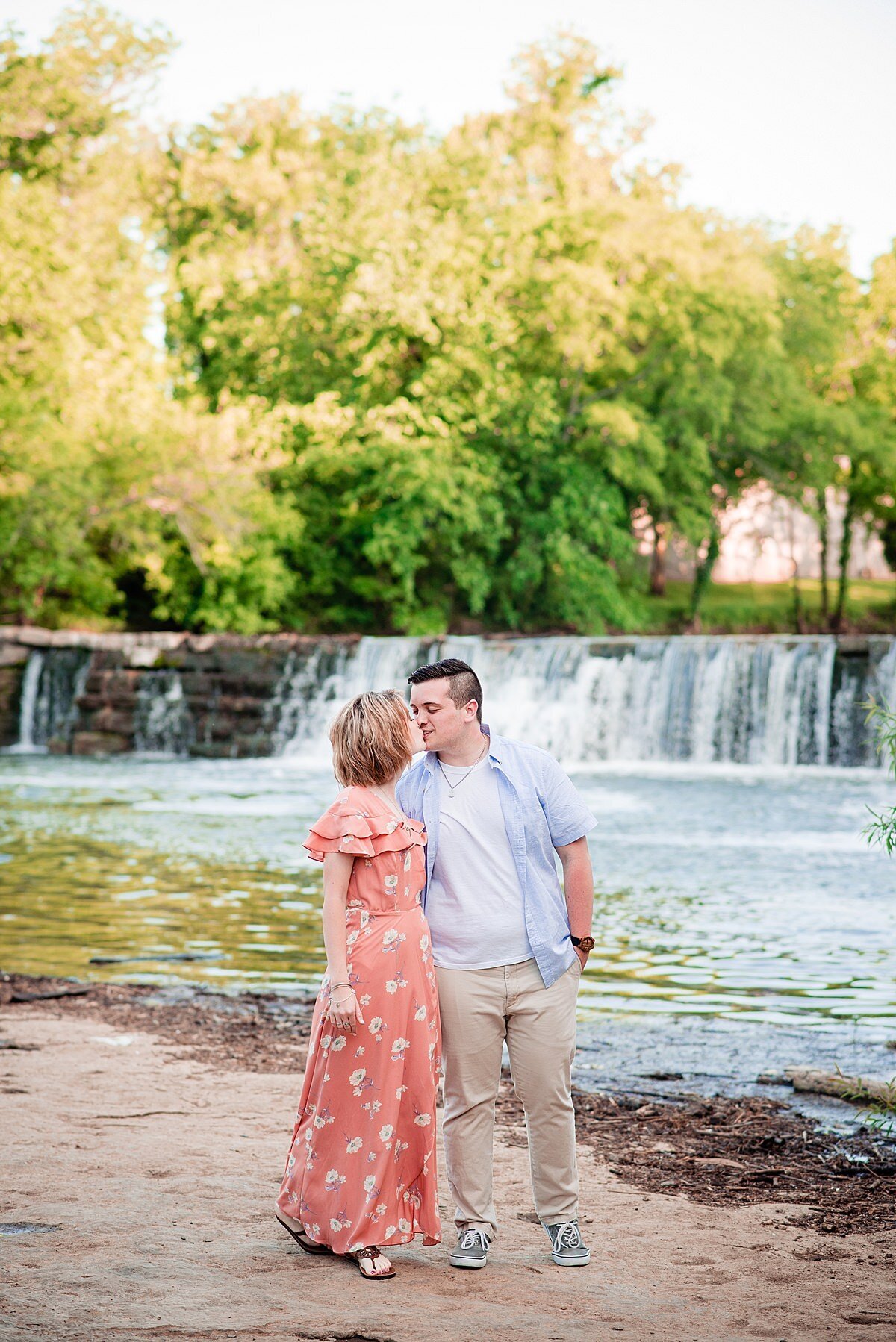 The bride and groom kiss in front of a waterfall. The groom has his arm around the bride's waist  and his hand in his pocket. He is wearing gray sneakers, khaki pants, a white t-shirt with a blue short sleeved shirt. The bride is wearing an off the shoulder floor length peach dress with a white floral pattern. She has two layers of ruffles around her shoulders.