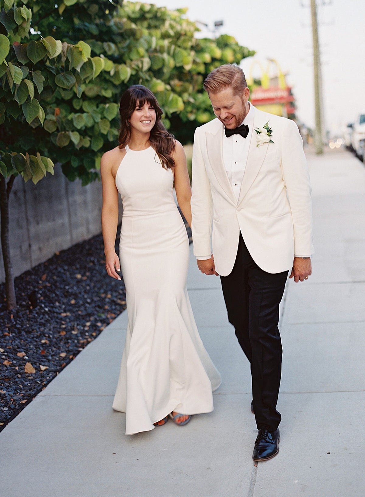 The bride, a brunette wearing a silk halter top sheath wedding dress, holds hand with the groom, who is wearing a tuxedo with a white jacket as they walk down the sidewalk in Nashville, Tennessee outside of Clementine Hall