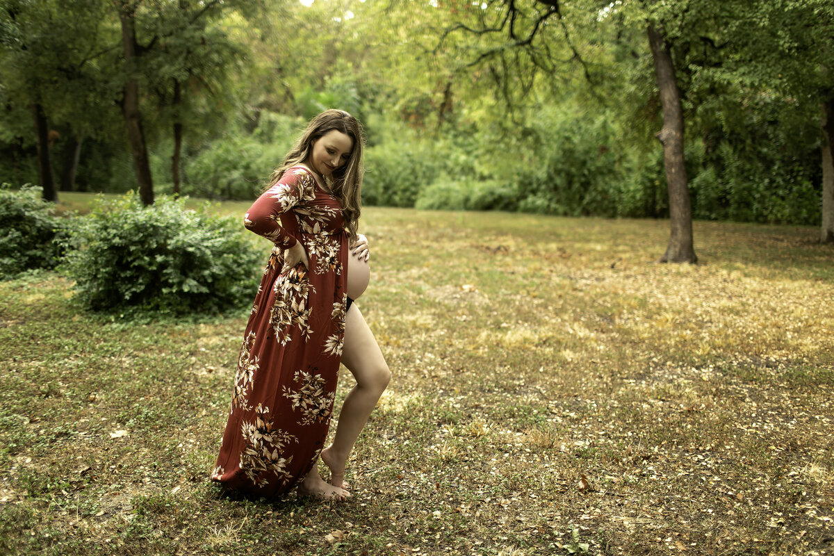 A serene maternity moment of the mom-to-be wearing a red dress, gazing lovingly at her belly while surrounded by the scenic beauty of Trinity Park, captured by Chunky Monkey Photography.