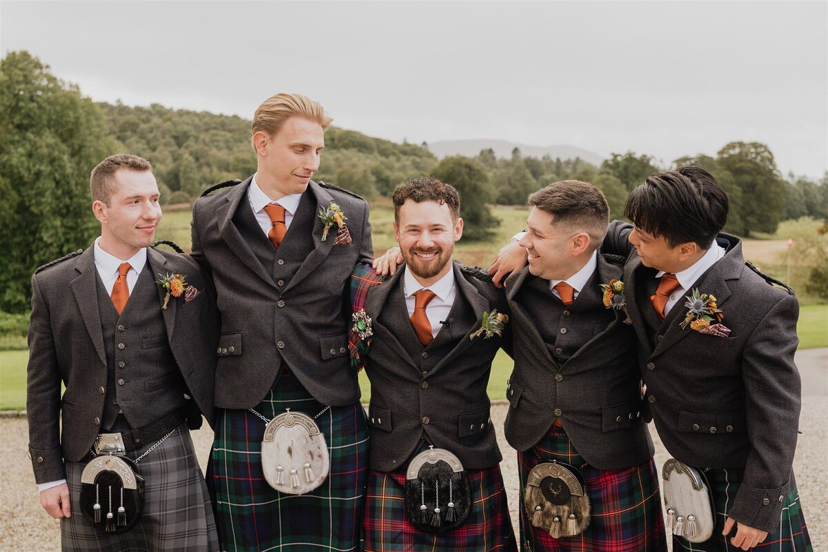 A wide photograph of a groom and his four groomsmen. They all hug each other in a line and smile candidly at the groom.