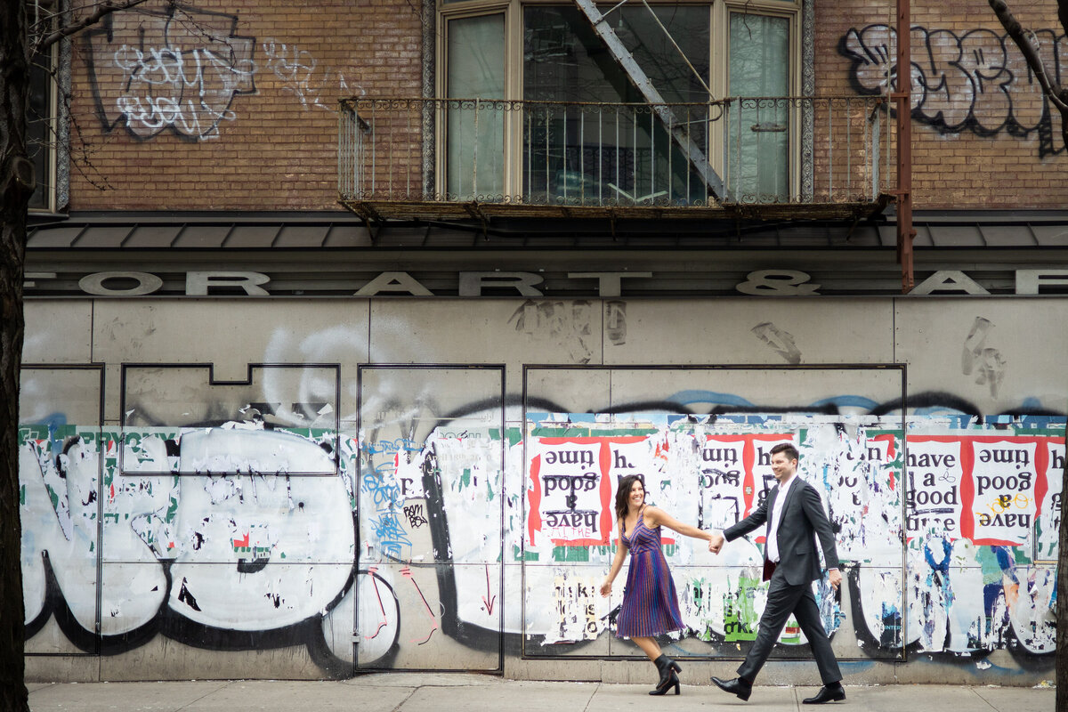 A couple holding hands and walking along a sidewalk in front of a graffiti covered wall.