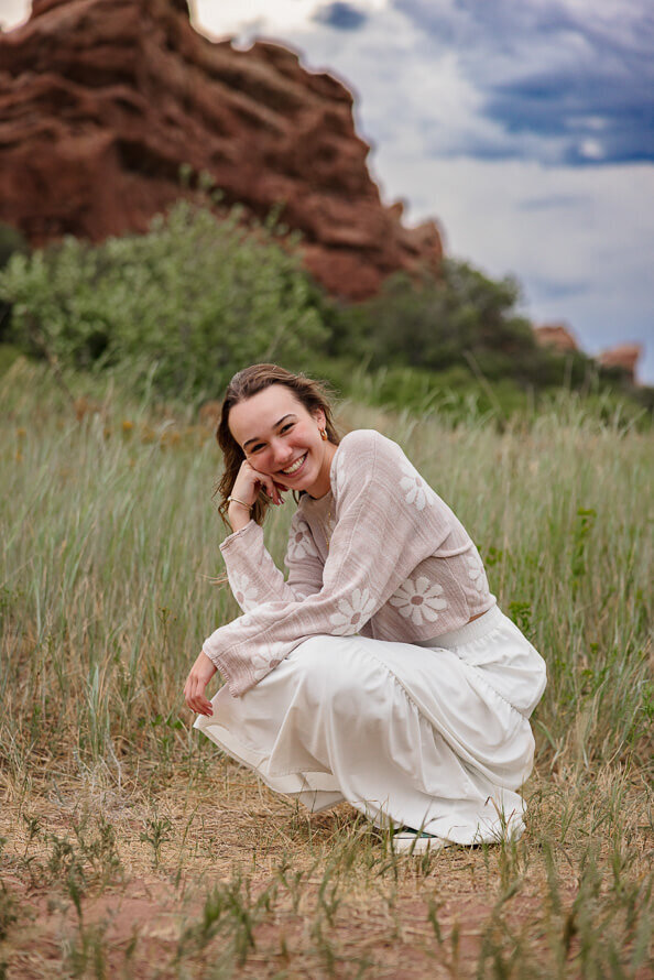 Girl squats and smiles at camera with red rocks behind her.