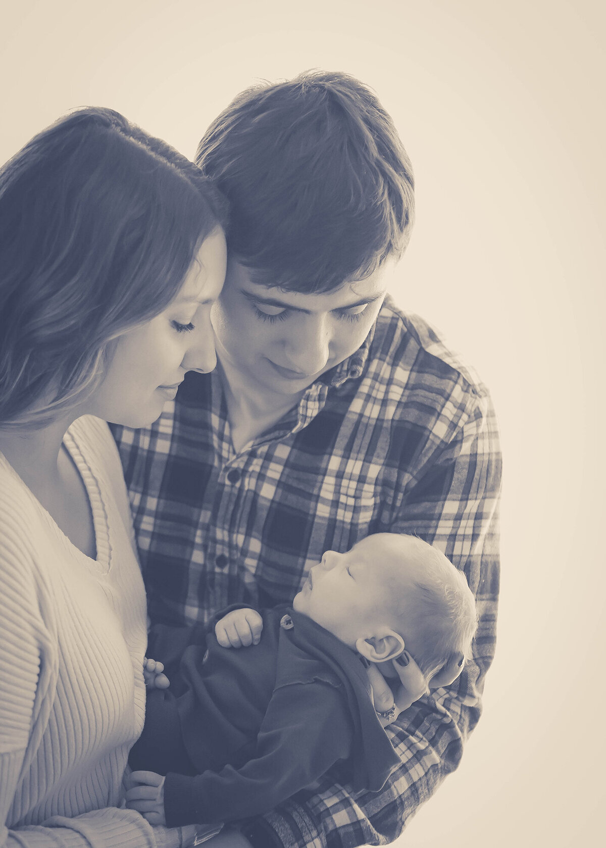 Happy new parents stand in the studio of an Iowa Newborn Photographer while smiling down at their newborn in mom's hands