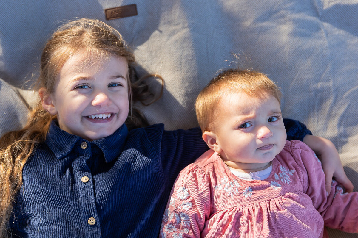 Two young children, a smiling girl in a blue shirt and a baby in a pink top, lying on a gray blanket outdoors.