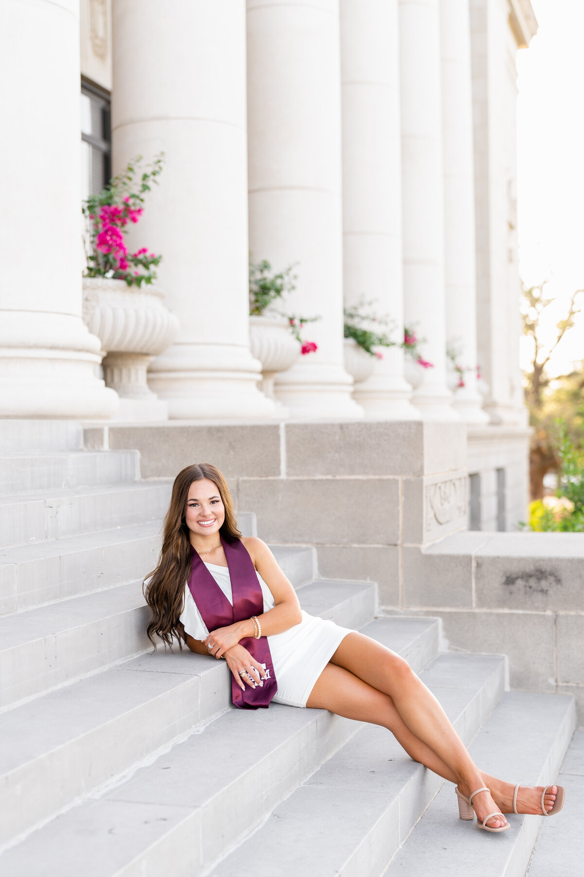 Texas A&M senior girl sitting on Administration Building stairs wearing white dress and maroon stole  while smiling