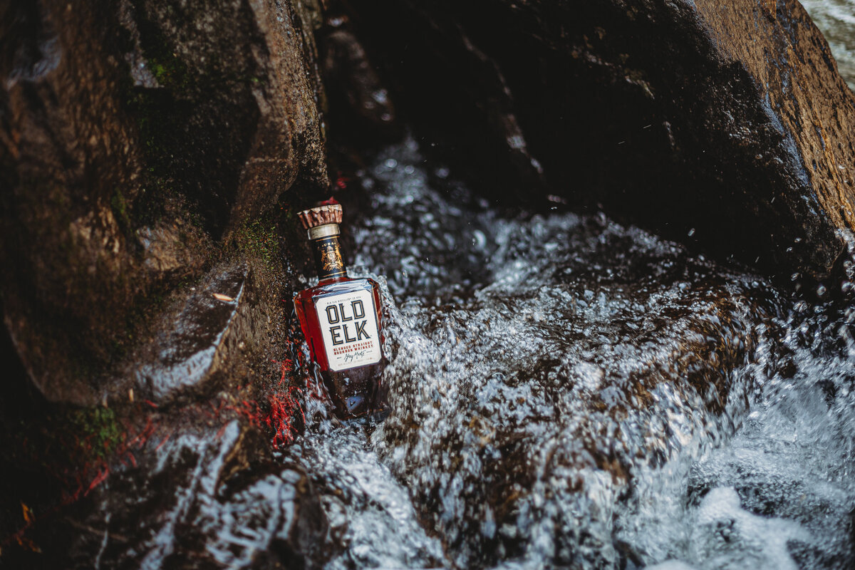 A bottle of Old Elk Whiskey sits in a river with water running past it.