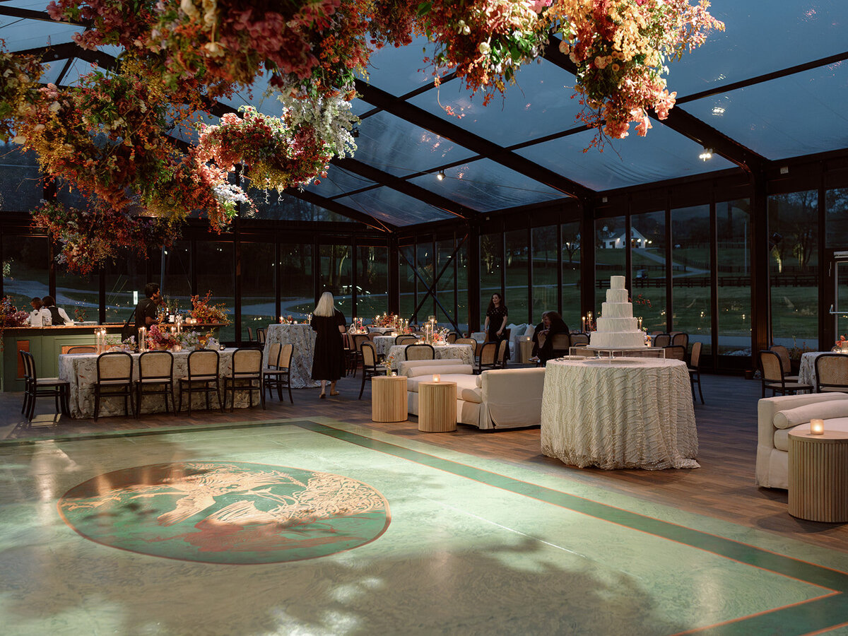 Interacting and sculptural floral clouds hang over the dance floor and bars of this fall wedding reception in Tennessee. Clear top tent build out for fall wedding reception in Tennessee hills.  Product clustered floral clouds in a tasteful fall color palette of copper, ruby, gold, burgundy, rust, and lavender. Orchids, bittersweet berries, butterfly ranunculus, and tulips are highlighted. Unique floral design with flower forward large scale installations. Design by Rosemary & Finch Floral Design in Nashville, TN.