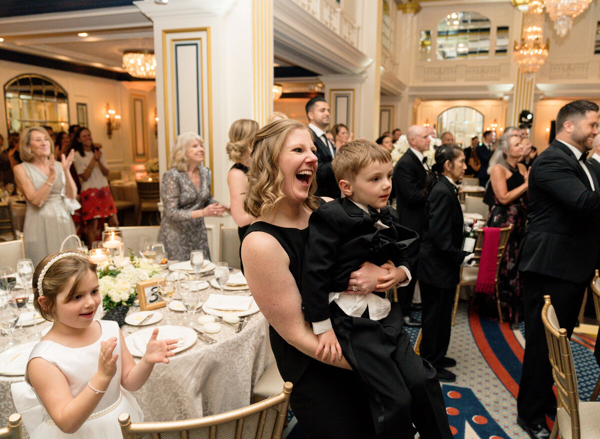 A joyful woman in a black dress holds a smiling young boy dressed in a suit at a formal event. A young girl in a white dress stands beside them clapping. The elegant room is filled with seated guests and decorative chandeliers.