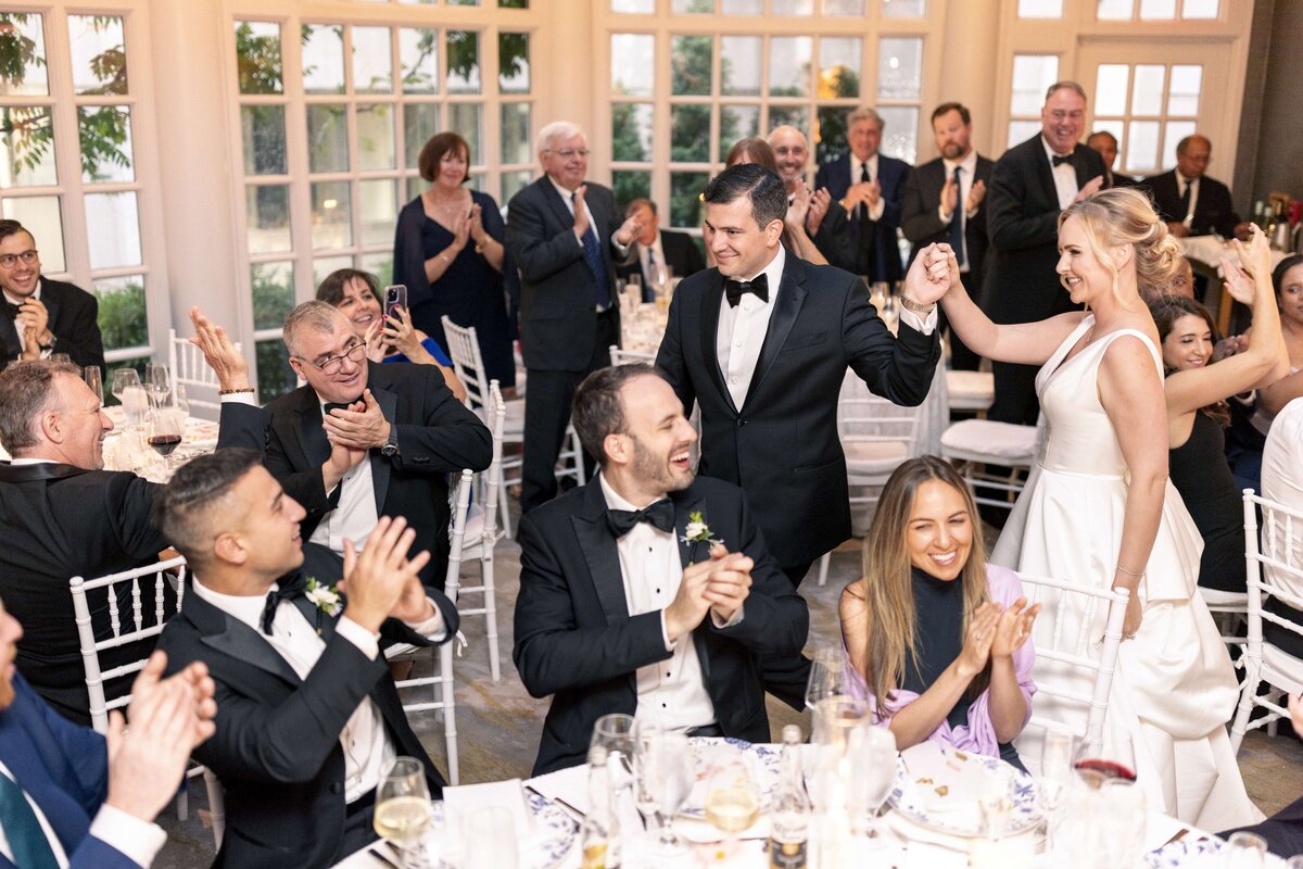A bride and groom, dressed elegantly, walk hand-in-hand through a reception area. Guests seated around tables clap and cheer. The atmosphere is celebratory, with warm lighting and large windows in the background.