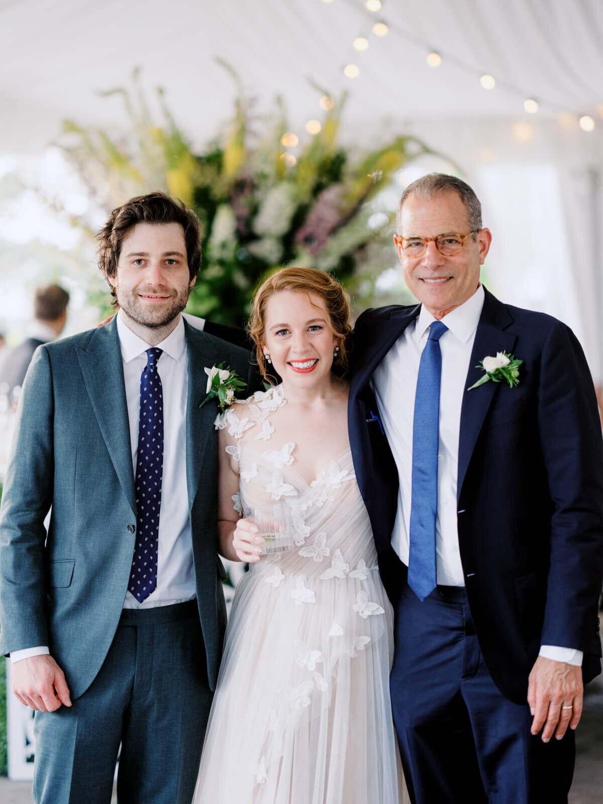 The bride and the groom, together with an old man in a suit, are standing and smiling on a large flower arrangement background.