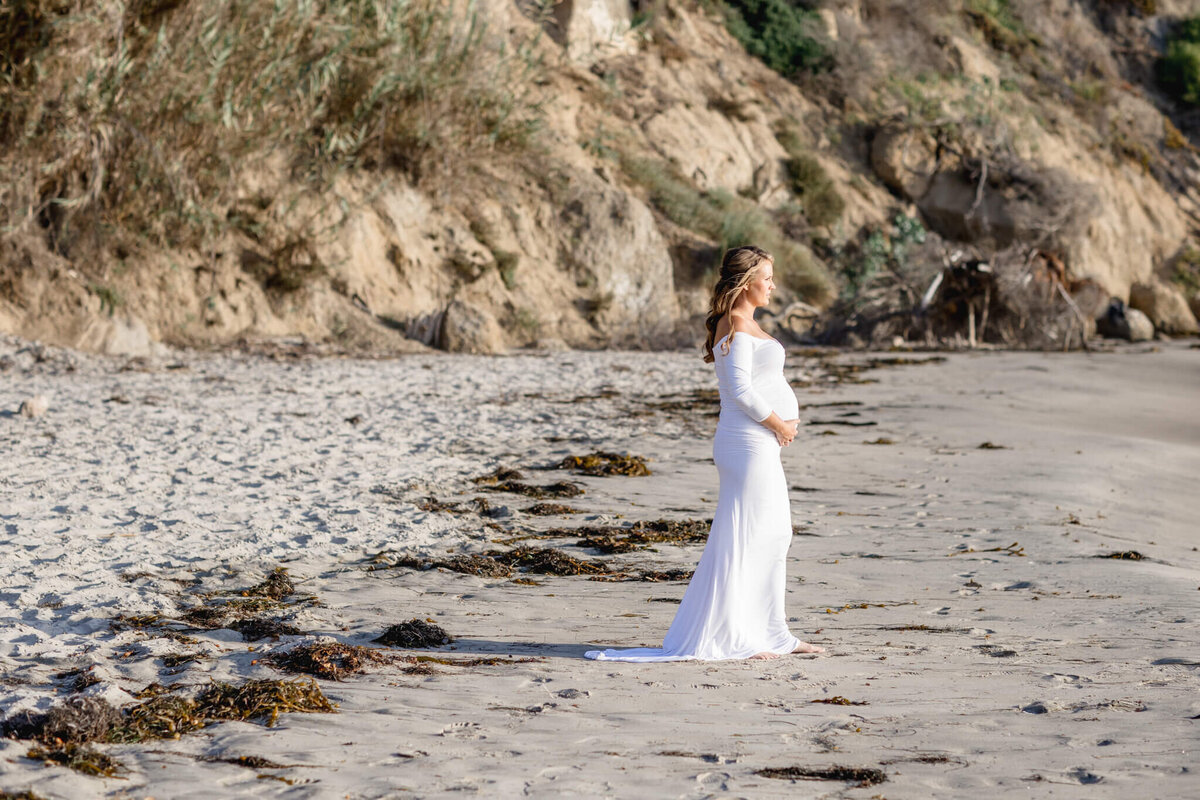 A woman in a long white dress stands on a sandy beach, looking out at the sea. She is barefoot, and the shoreline is lined with rocks and vegetation. The sunlight casts shadows, enhancing the serene atmosphere.