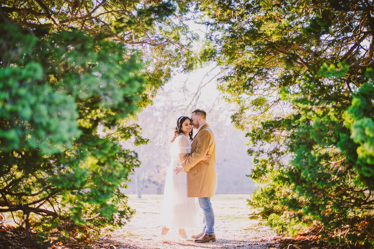 A couple standing under a tree tunnel at the planting fields arboretum in the fall during their engagement session