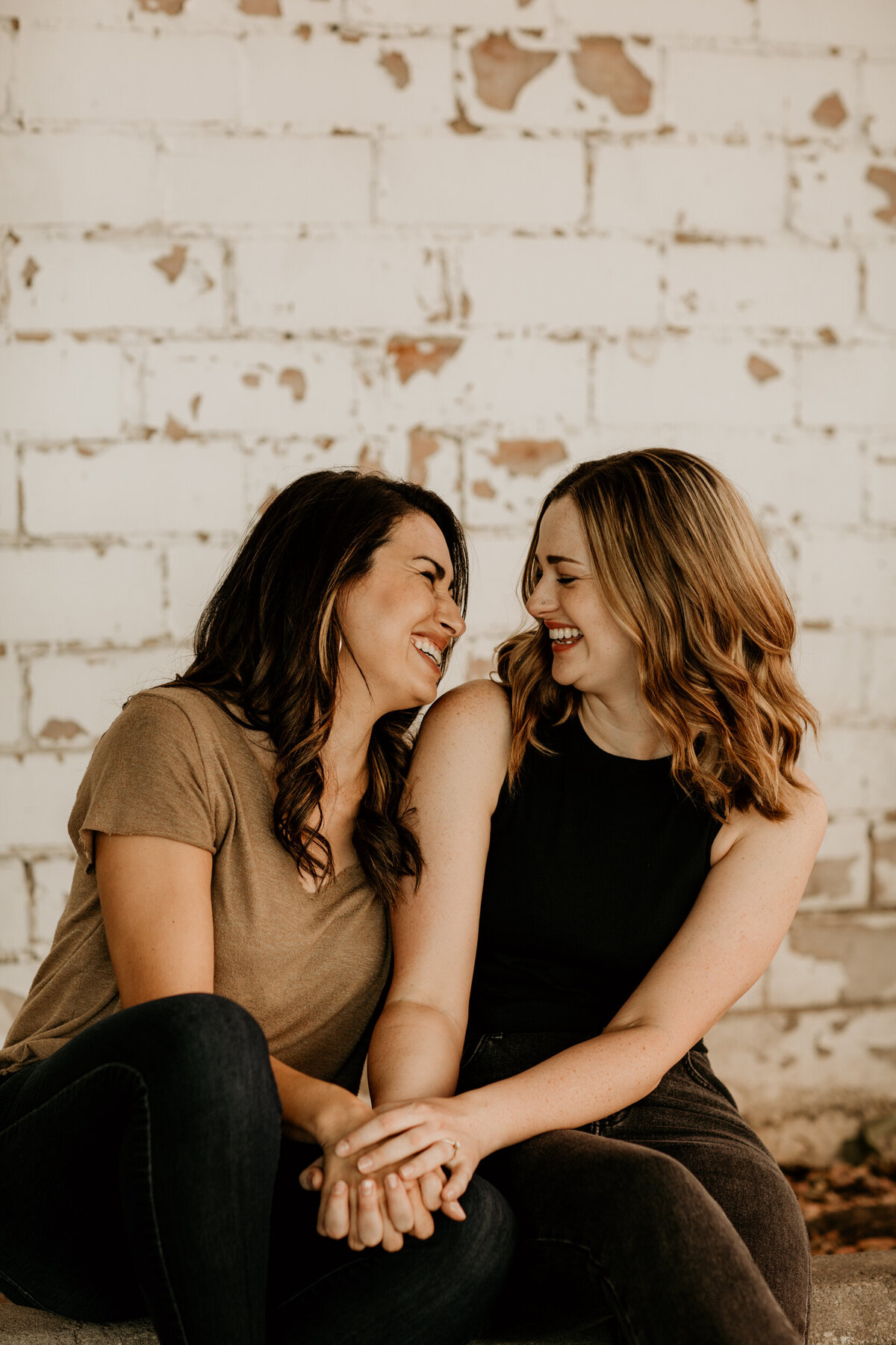 same sex couple laughing  and sitting together in front of a white brick wall