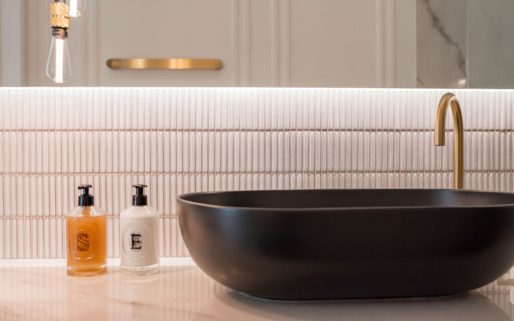 Modern bathroom with marble countertops and white tile backsplash. A black vessel sink with gold hardware sits on the counter.