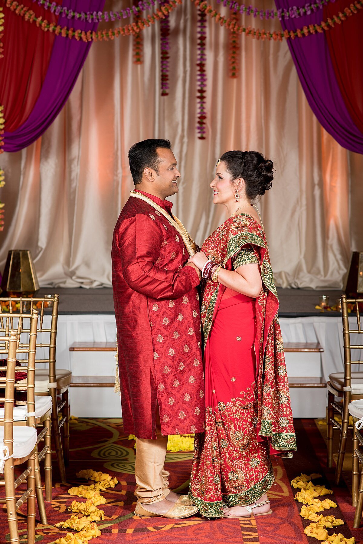 Hindu groom wearing a gold and red sherwani and an Indian bride wearing a red, green and gold saree holding hands in front of their magenta, peach and red mandap decorated with floral garlands at their Nashville wedding