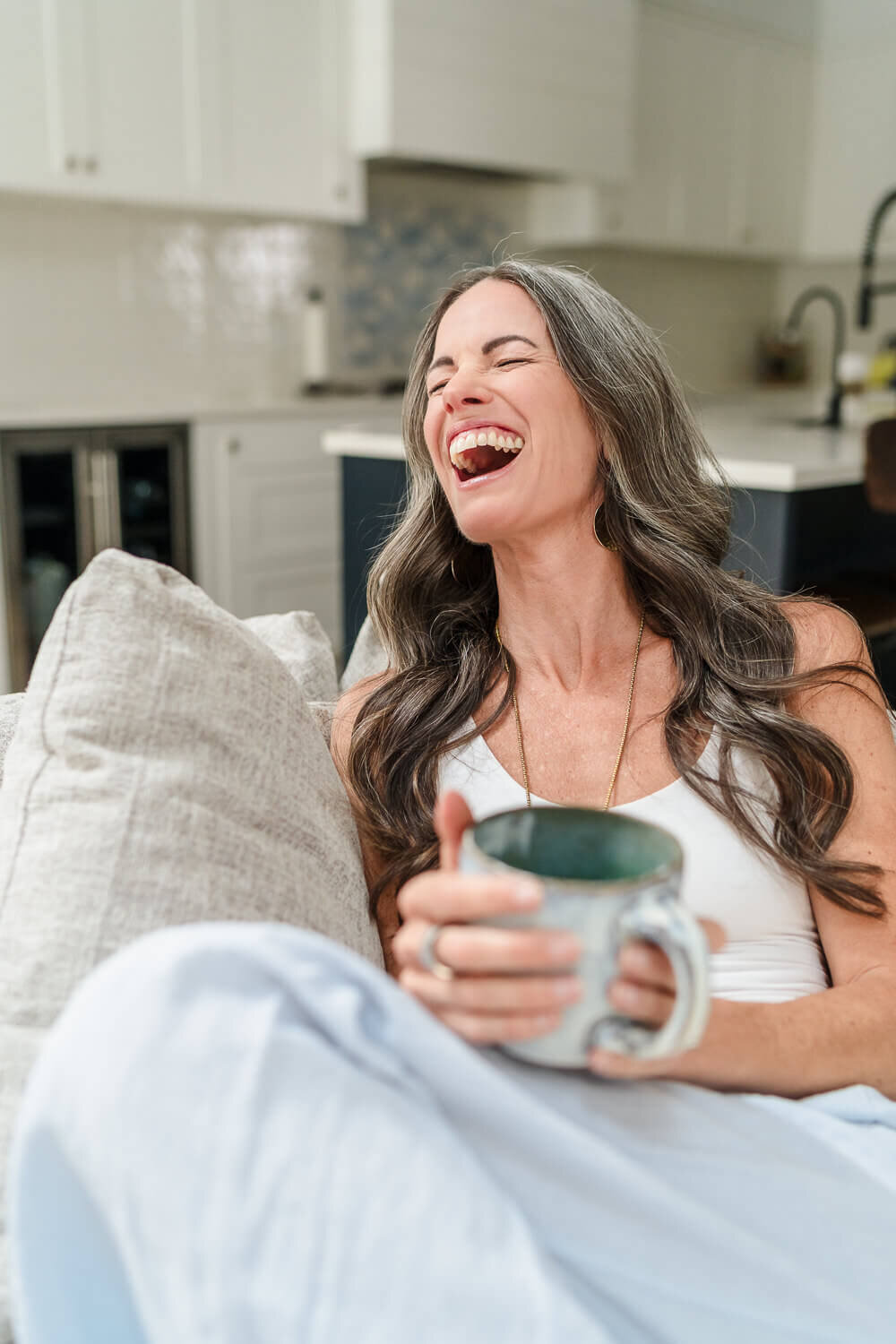 Jenn Pike laughing on couch in white tank top, holding speckled ceramic mug in modern kitchen.