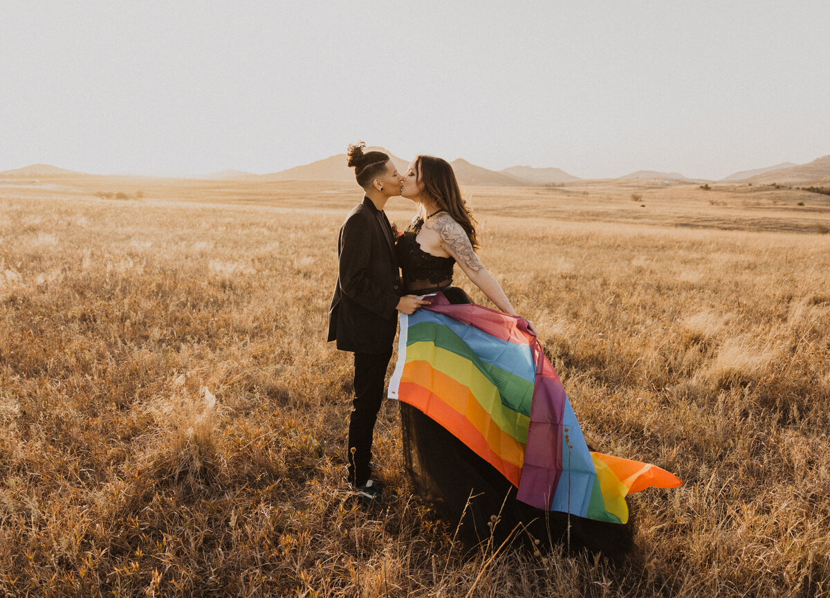 An lgbtq elopement in the wichita mountains in oklahoma. The couple is holding a rainbow flag dressed in untraditional black wedding clothes during golden hour