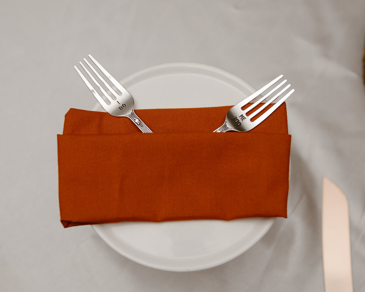 Overhead view of two vintage silver forks engraved with “I DO” and “ME TOO,” resting on a rust-colored napkin over a white plate at a wedding reception table.